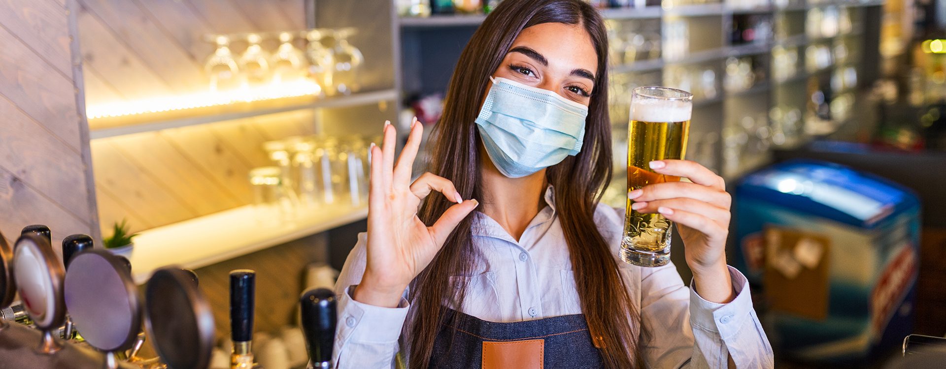 Bartender with protective face mask,covid-19 protection, serving a draft beer at the bar counter during coronavirus pandemic, showing OK sign,shelves full of bottles with alcohol on the background